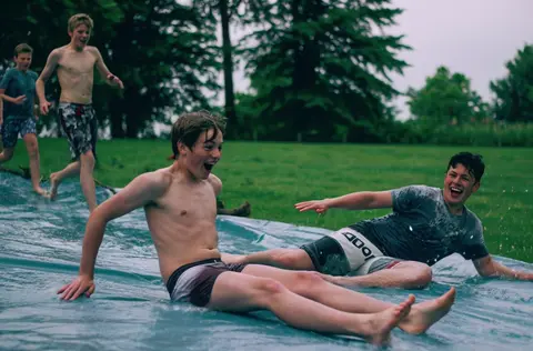 Grok Imagine 1.5 clip: teenage boys sliding down a wet plastic tarp on a lawn