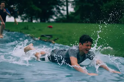 PixVerse V6 clip: teenage boys sliding down a wet plastic tarp on a lawn